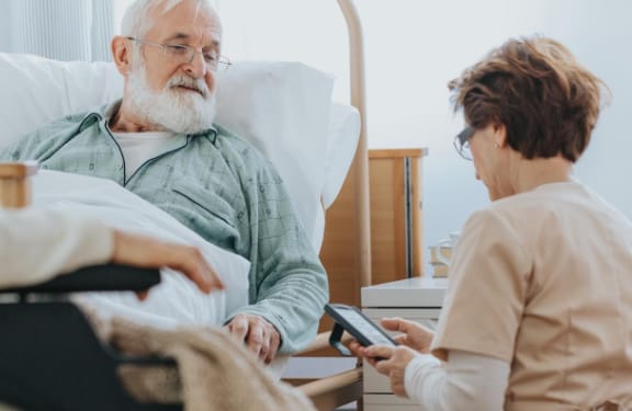 Caregiver kneeling down beside hospital bed asking an elderly man some questions